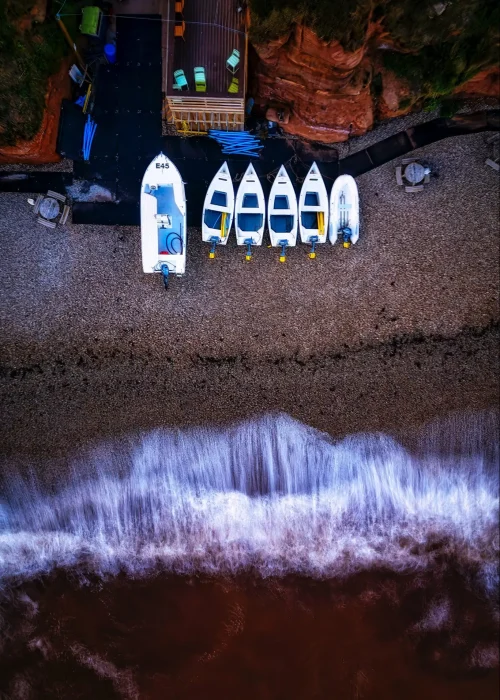 aerial view of waves crashing against beach and red cliffs with boats along the sand in Tavin